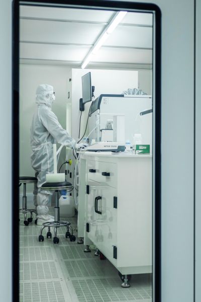 a technician in a full white sterile cleanroom suit stands at a white laboratory bench operating testing equipment inside a brightly lit, sterile cleanroom environment